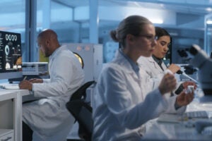 Three scientists in lab coats work in a modern laboratory; one uses a computer, another examines samples, and the third looks into a microscope. The setting is bright with scientific equipment.
