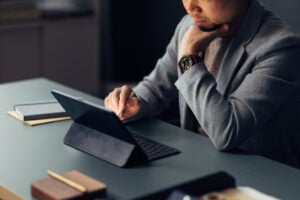 A person in a blazer sits at a desk, using a tablet with a keyboard case. They are resting their chin on one hand and appear to be focused on the screen. A notebook and pen are also on the desk.