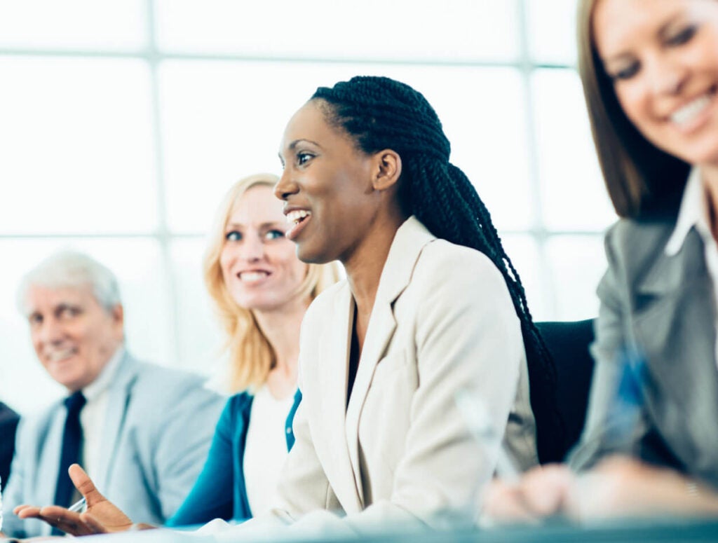 Four professionals sit in a row at a meeting table. The focus is on a woman in a light blazer speaking and smiling, while the others listen and smile. The background is bright and out of focus.