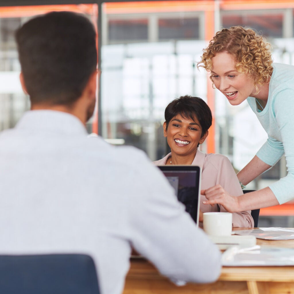 Three colleagues collaborate in a modern office. One woman stands, smiling and pointing at a laptop screen, while another woman and a man sit at the table, engaged and cheerful. A coffee cup and papers are on the table.