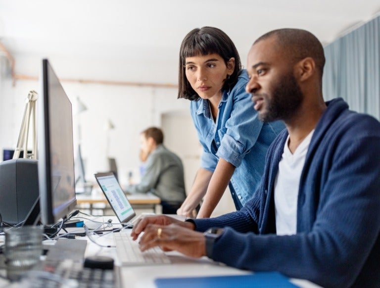 Two coworkers in a modern office work together at a computer. One person sits and types on the keyboard, while the other stands beside, leaning in and looking at the screen. Other workers are in the background.
