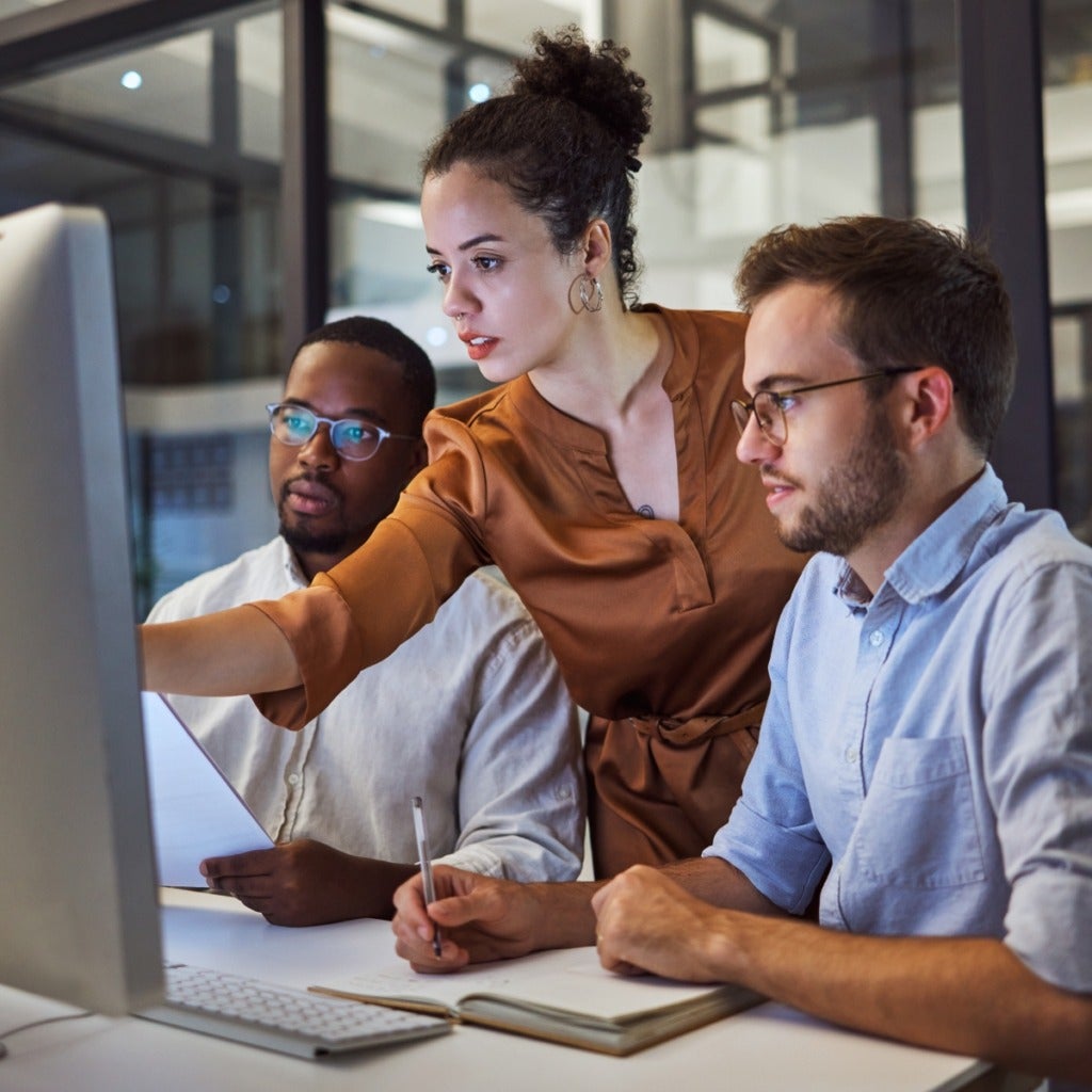 Three co-workers at a desk, having a discussion while looking at a large computer monitor.